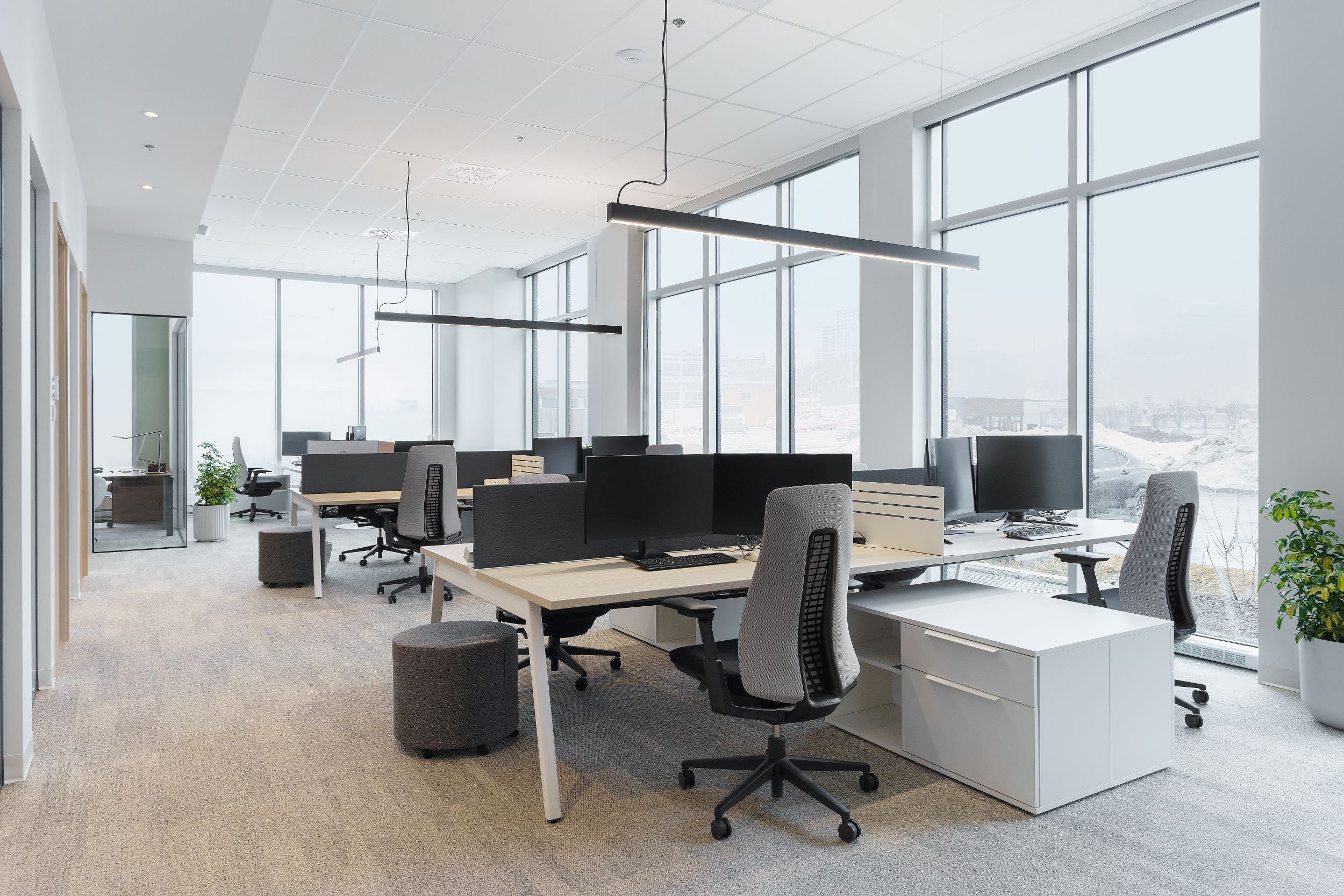 Espace de travail à aire ouverte moderne avec de grandes fenêtres, postes de travail partagés équipés de chaises ergonomiques grises, bureaux blancs et séparateurs noirs, éclairés par une suspension linéaire noire