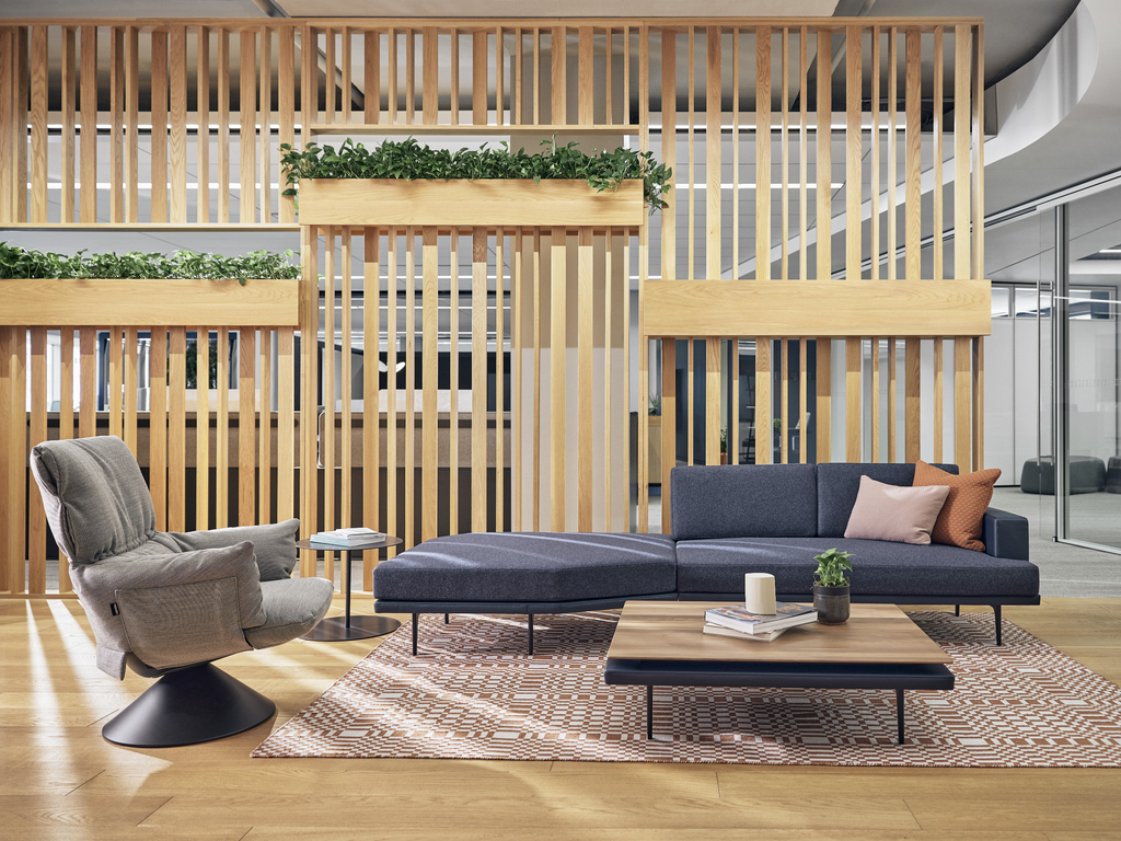 Espace lounge de bureau moderne avec fauteuil ergonomique, canapé design, cloison en bois décorative et table basse élégante.