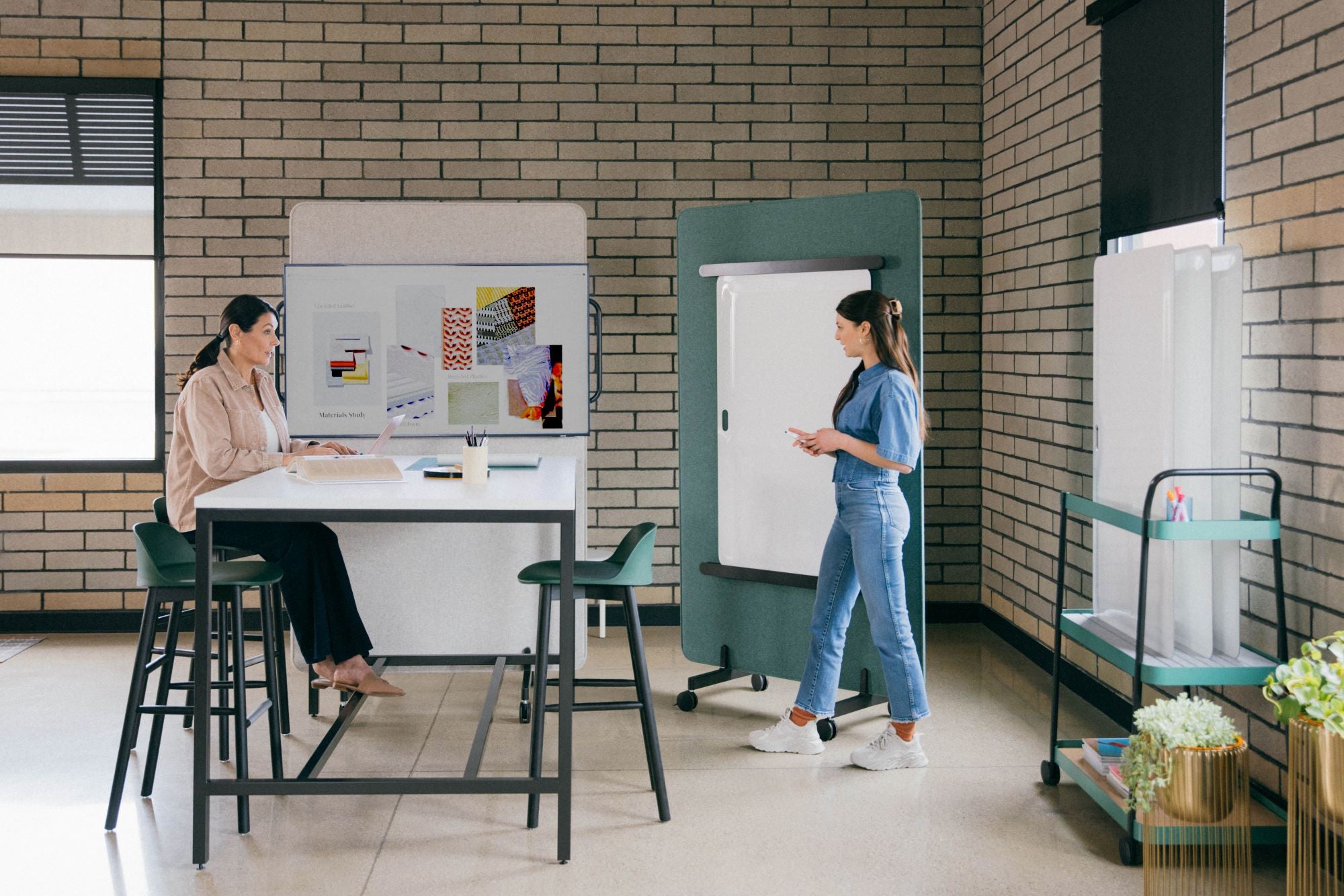 Salle de réunion collaborative avec table haute, tableau mobile et mobilier moderne. Deux collègues échangent autour d’un espace de travail créatif et flexible.