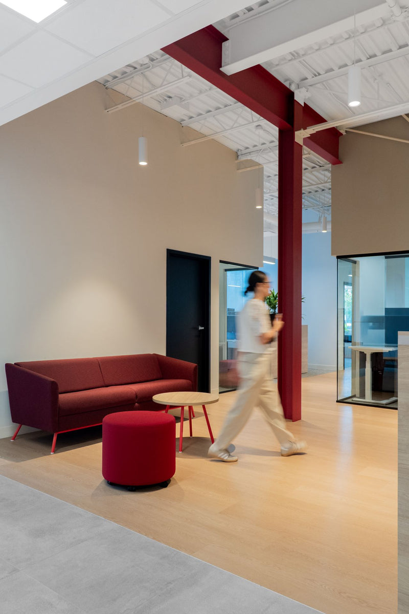 Espace lounge ouvert avec canapé bordeaux, pouf rouge et table basse en bois — employée en mouvement, colonnes métalliques rouges apparentes — Beauce Atlas