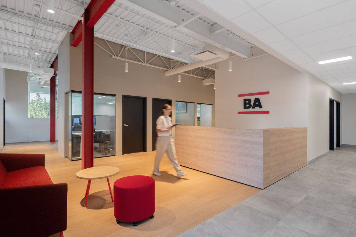 Réception Beauce Atlas avec comptoir en bois clair, logo BA, pouf rouge et canapé bordeaux, colonnes métalliques rouges apparentes