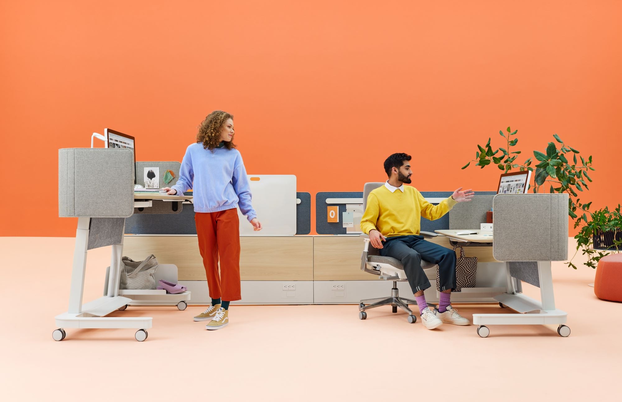 Espace de travail moderne avec bureaux réglables en hauteur, cloisons acoustiques et design ergonomique. Deux collègues collaborent dans un bureau lumineux et coloré.