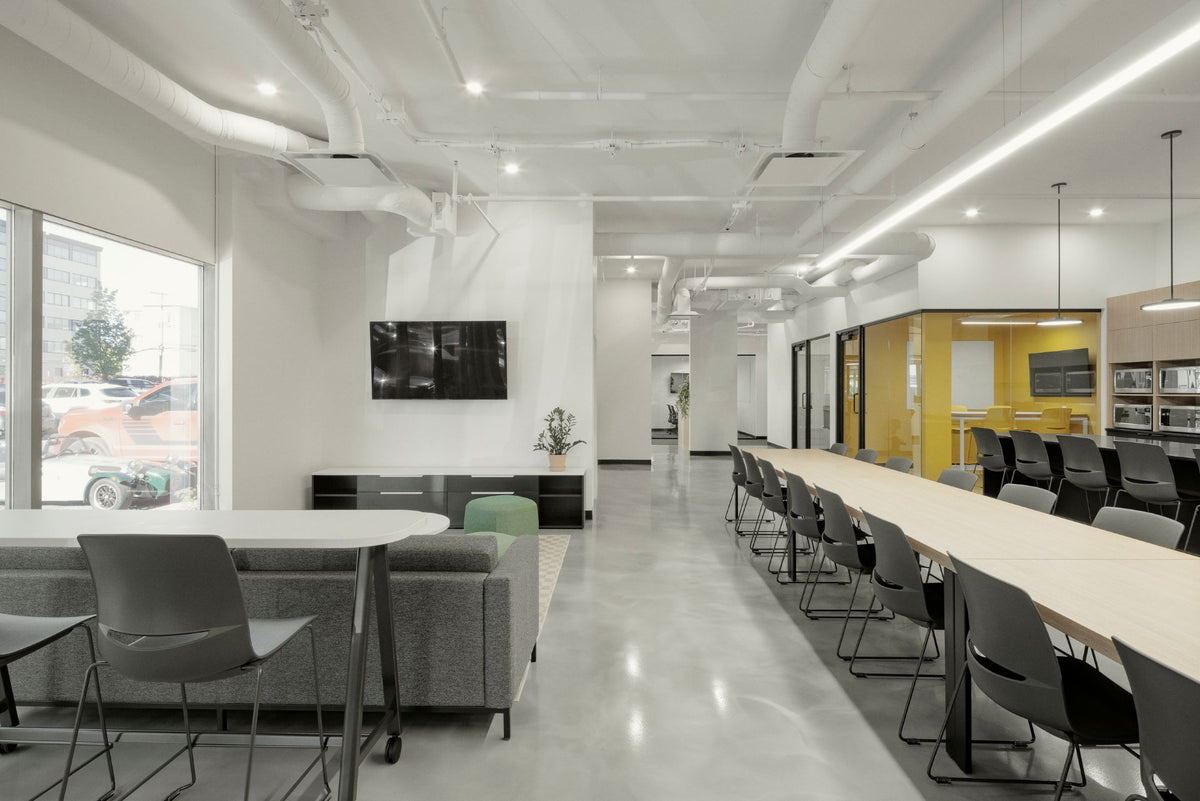 Espace cafétéria corporatif moderne avec grande table communautaire, coin détente avec écran mural et mobilier contemporain aux accents colorés. Aménagement lumineux à aire ouverte incluant tables hautes, chaises design et cuisinette intégrée pour créer un environnement de travail collaboratif et convivial.