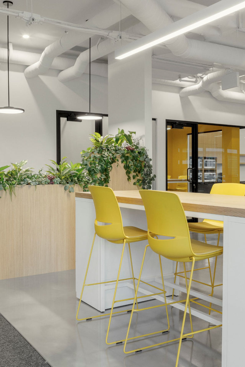 Îlot de travail collaboratif moderne avec tabourets hauts jaunes, jardinières intégrées et design biophilique pour espaces de bureau contemporains. Aménagement d'entrée accueillant combinant fonctionnalité et esthétique avec plafond industriel apparent, éclairage LED et accents de couleur dynamiques.