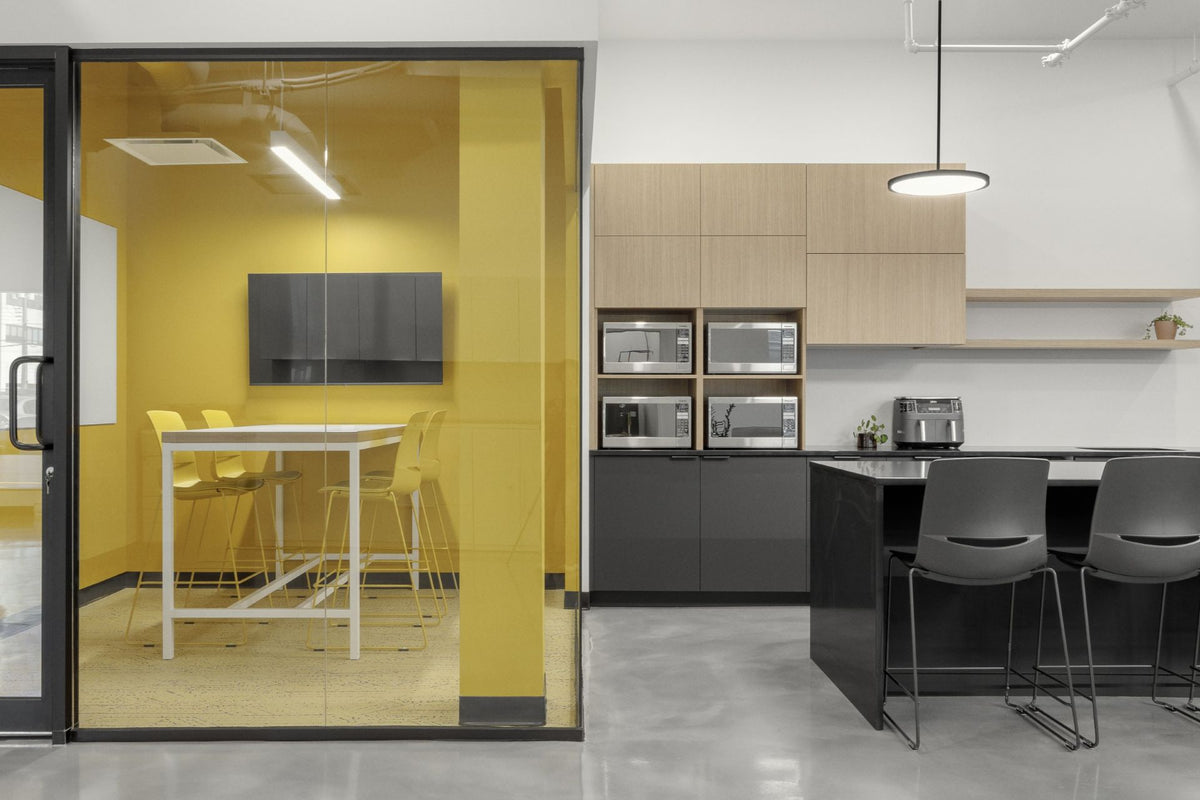 Salle collaborative vitrée avec murs jaunes vibrants, table haute blanche et chaises design, adjacente à une cuisinette corporative moderne avec armoires grises et îlot noir. Espace de travail d'équipe dynamique combinant zone de réunion privée et coin repas avec électroménagers intégrés pour environnement de bureau contemporain.