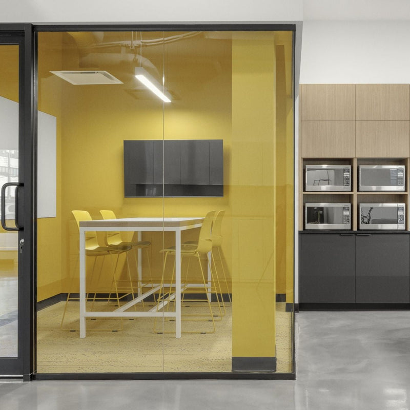 Salle collaborative vitrée avec murs jaunes vibrants, table haute blanche et chaises design, adjacente à une cuisinette corporative moderne avec armoires grises et îlot noir. Espace de travail d'équipe dynamique combinant zone de réunion privée et coin repas avec électroménagers intégrés pour environnement de bureau contemporain.