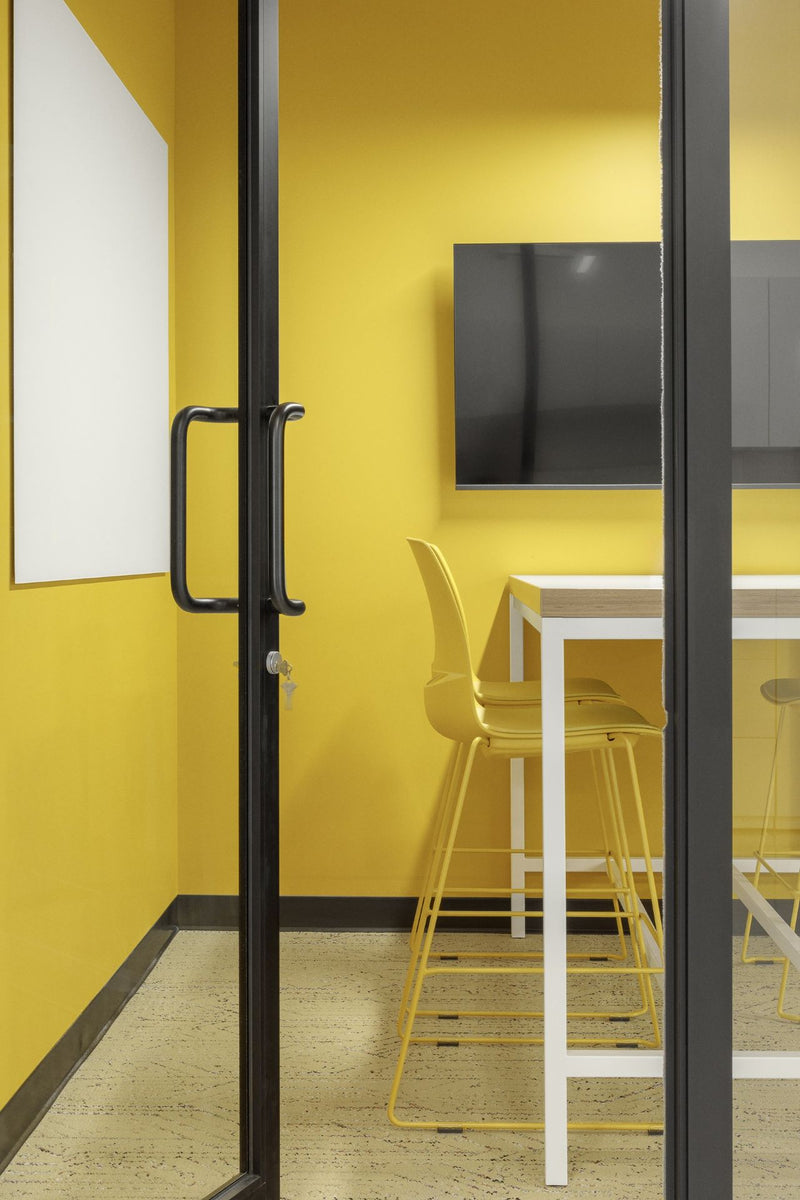 Salle de réunion collaborative avec murs jaunes énergisants, porte vitrée à cadre noir et mobilier minimaliste incluant table haute blanche et tabourets jaunes assortis. Espace de travail d'équipe moderne avec écran mural gris pour présentations, design audacieux et ambiance stimulante pour favoriser la créativité et la productivité.