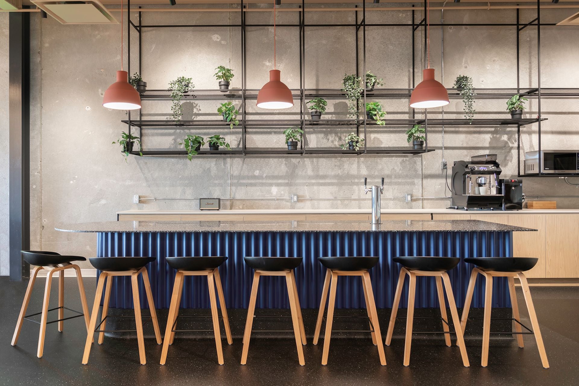 Cuisine de bureau moderne avec îlot central bleu cannelé et tabourets en bois - espace collaboratif professionnel avec suspensions design et étagères végétales