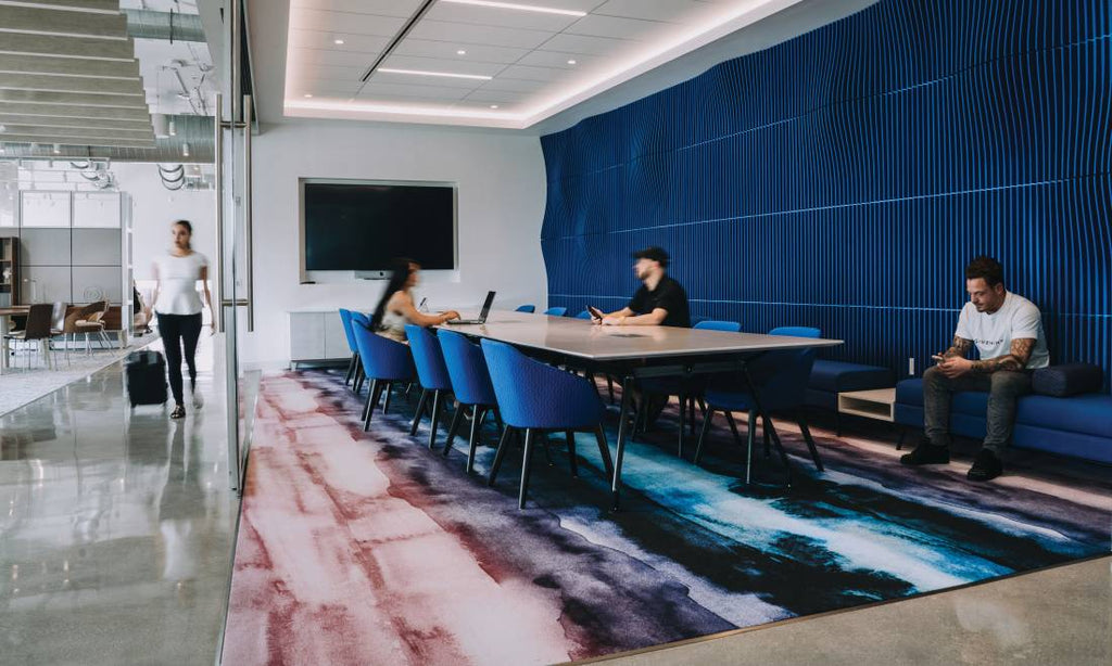 Salle de réunion moderne avec grande table, chaises bleues design, mur décoratif texturé et tapis coloré contemporain.